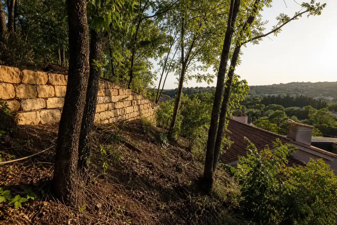 Parcelle en pente boisée sur un coteau de l’ouest lyonnais, mur de soutènement en pierre et maisons en contrebas